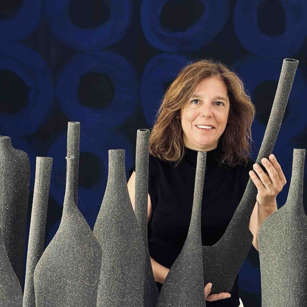 Retrato de Sofia Aguas, sorridente com cabelo castanho médio, vestindo uma blusa preta sem mangas, posicionada entre várias esculturas de cerâmica de textura áspera e formato alongado, semelhantes a garrafas ou vasos. O fundo apresenta um padrão abstrato em tons de azul escuro, criando um contraste elegante com as esculturas cinza.