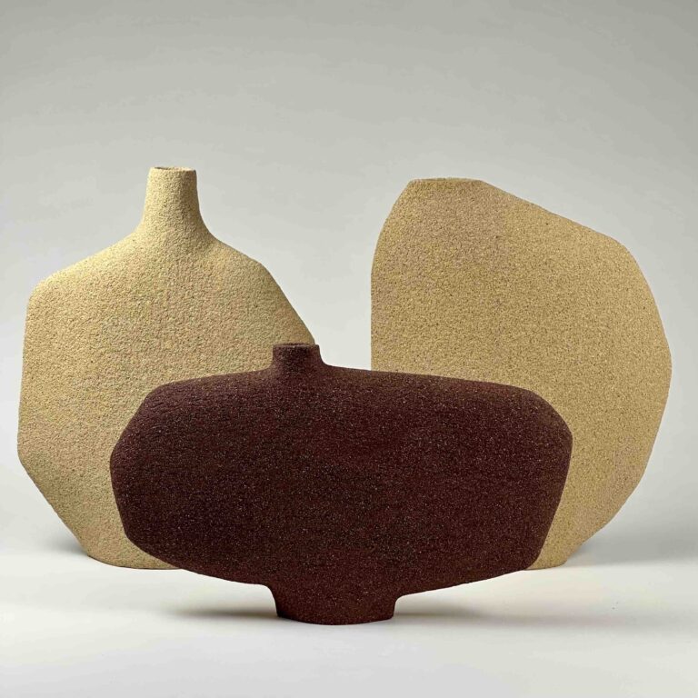 três vasos de cerâmica de formato contemporâneo e assimétrico. Cada um possui uma textura áspera e tons terrosos em amarelo e vermelho dispostos em um fundo neutro.