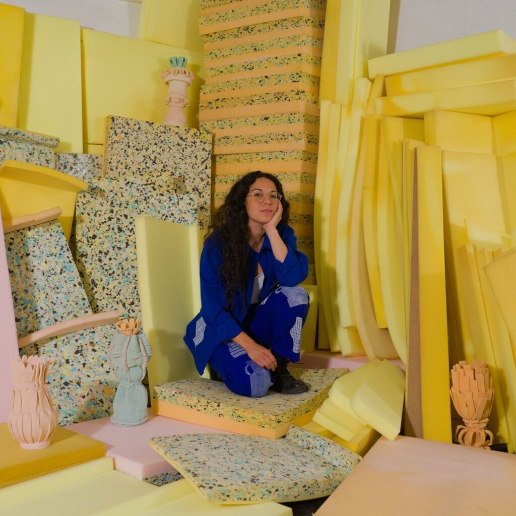 A artista Margarida lopes pereira sentada no seu estúdio, rodeada por blocos de espuma amarela e peças de espuma. O ambiente vibrante e lúdico reflete o universo criativo e experimental do seu trabalho.