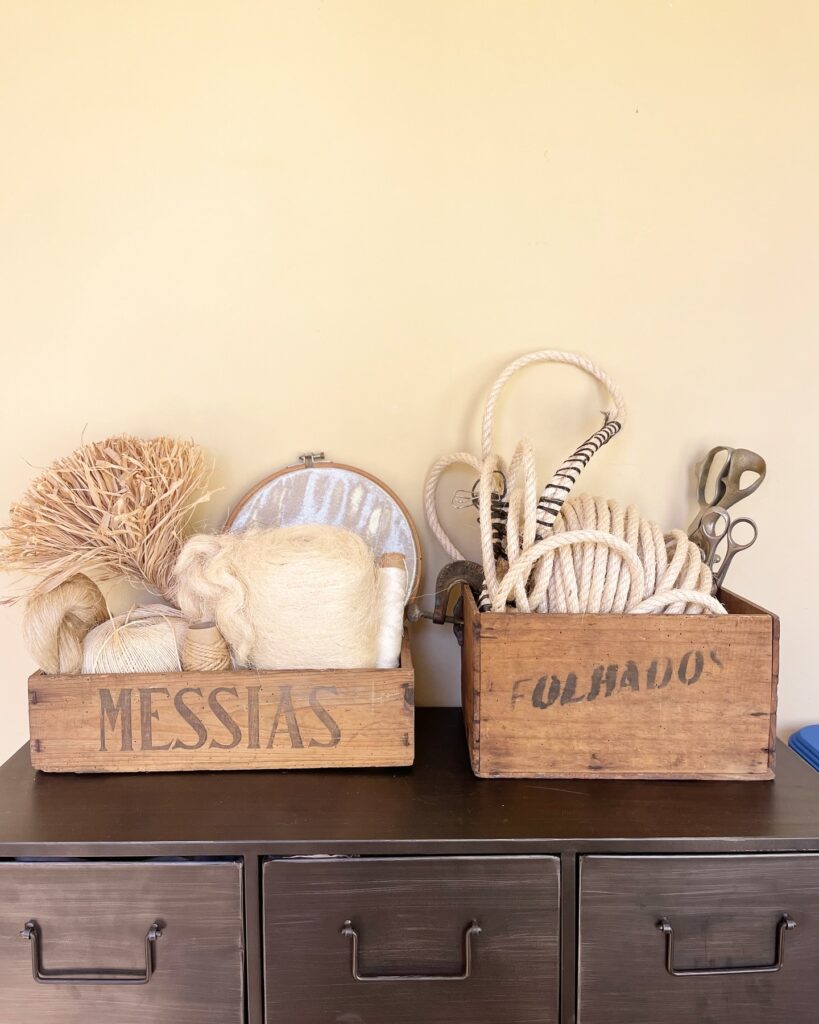 Caixa de madeira com fios de lã, fibras naturais e materiais têxteis para escultura. Ao lado, outra caixa com cordas e ferramentas artesanais no atelier de Maria Pratas.