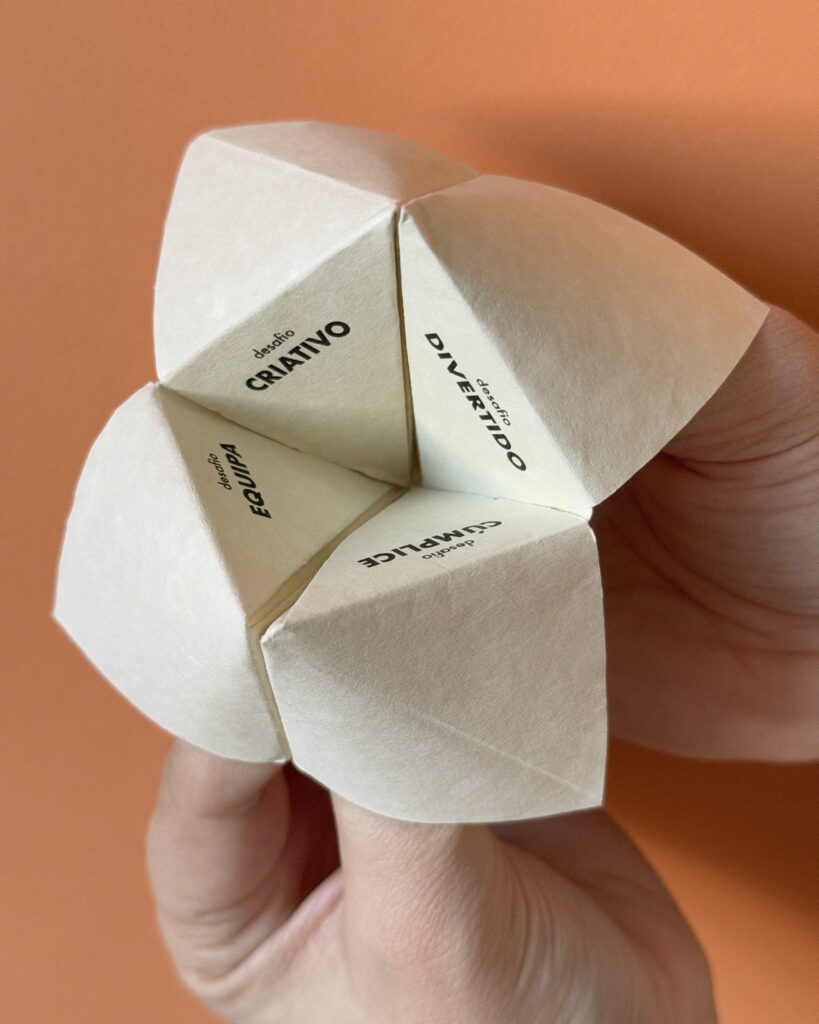 Mãos segurando um jogo de origami “Quantos-Queres” feito de papel branco, com palavras como “Criativo”, “Divertido”, “Equipa” e “Cúmplice” escritas em cada seção, sobre um fundo laranja.