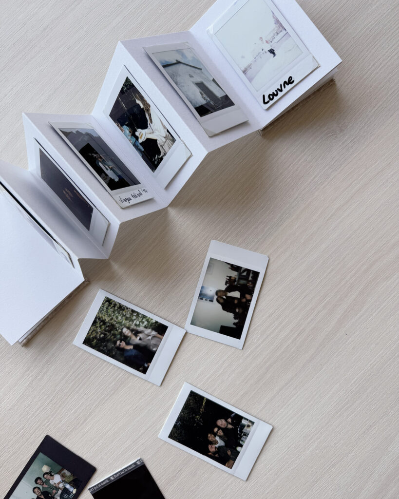 Álbum sanfonado aberto sobre uma mesa clara, exibindo fotos instantâneas estilo polaroid. Algumas imagens estão espalhadas ao redor, capturando momentos especiais entre amigos e familiares.