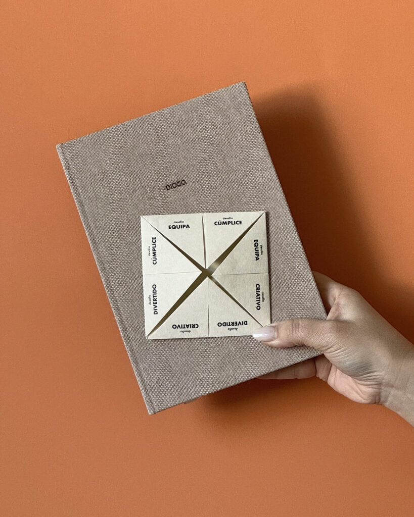Mão segurando um livro de capa dura com textura bege, sobre um fundo laranja. Sobre o livro, há um jogo de origami “Quantos-Queres” em papel, com palavras como “Cúmplice”, “Criativo”, “Divertido” e “Equipa” impressas em cada seção.