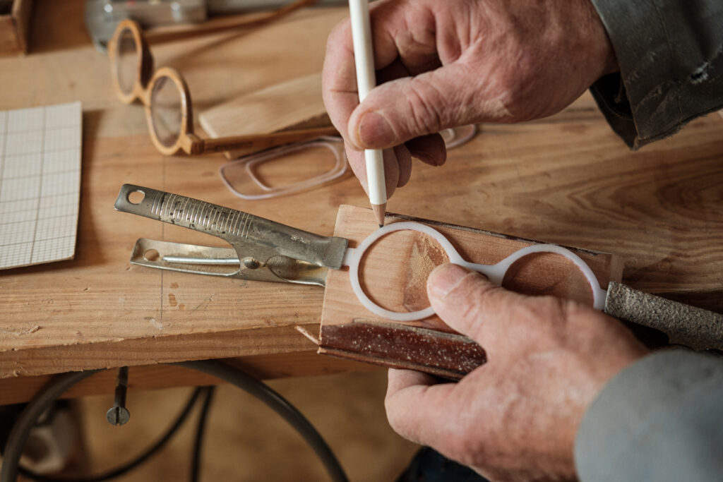 Artesão traçando o contorno de uma armação de óculos em madeira, sobre uma bancada de trabalho com ferramentas e materiais artesanais ao redor.
