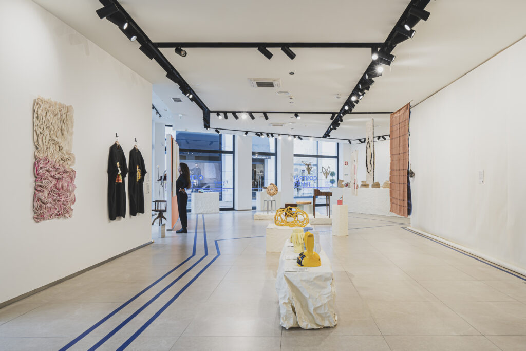 O espaço apresenta um design minimalista e contemporâneo, com peças artesanais dispostas sobre pedestais de papel enrugado e suspensas nas paredes. No primeiro plano, esculturas e objetos de design artesanal em materiais como cerâmica e fibra natural. À esquerda, um têxtil artístico em tons de rosa e bege, ao lado de duas vestes pretas penduradas na parede. No fundo, uma visitante observa uma das obras expostas. A iluminação realça os detalhes das peças e cria uma atmosfera imersiva.