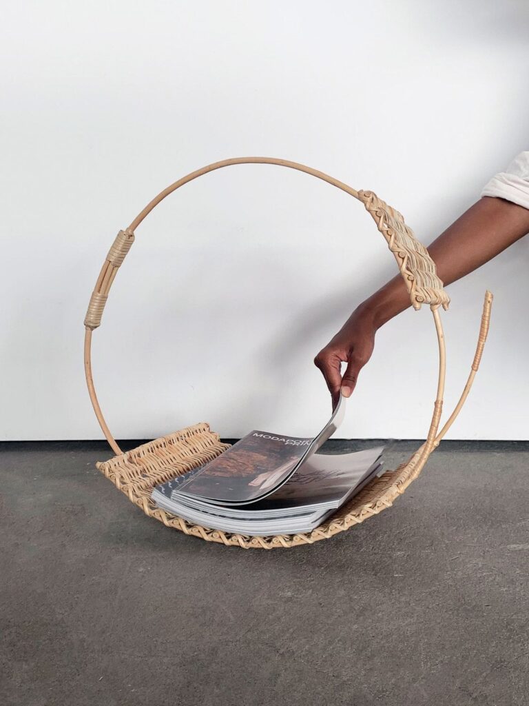 Mão a retirar uma revista de um suporte circular de vime. A peça apresenta um design inovador, reinterpretando a cestaria tradicional com formas minimalistas e estruturais.