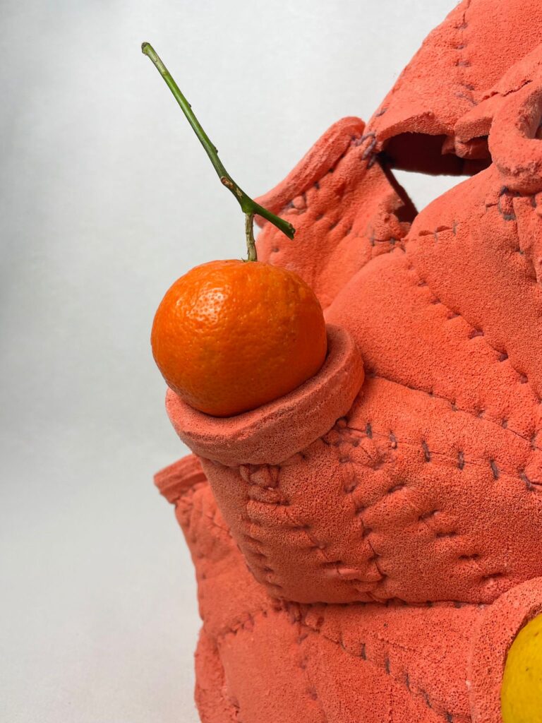 Detalhe de uma peça de espuma texturizada em tom terracota, com costuras aparentes, segurando uma laranja com caule dentro de uma pequena cavidade. O design artesanal explora forma e textura de maneira orgânica.