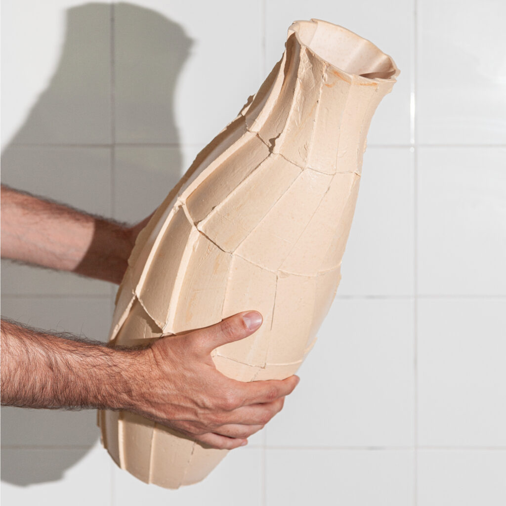 Mãos seguram uma jarra de cerâmica com superfície facetada e acabamento cru. A peça tem um design escultural e experimental, destacando as texturas e imperfeições do material.
