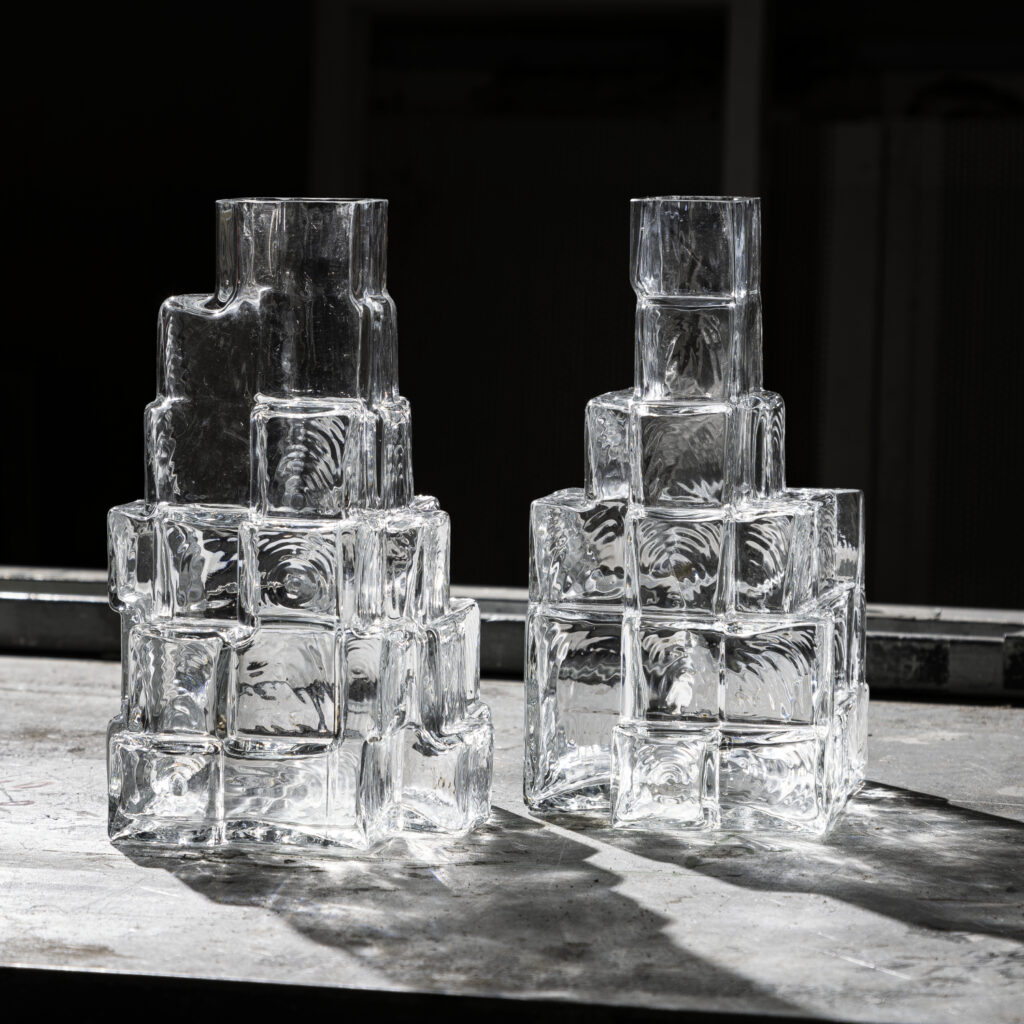 Duas jarras de vidro empilhado com forma geométrica, inspiradas em cubos sobrepostos, refletindo a luz de forma única.
