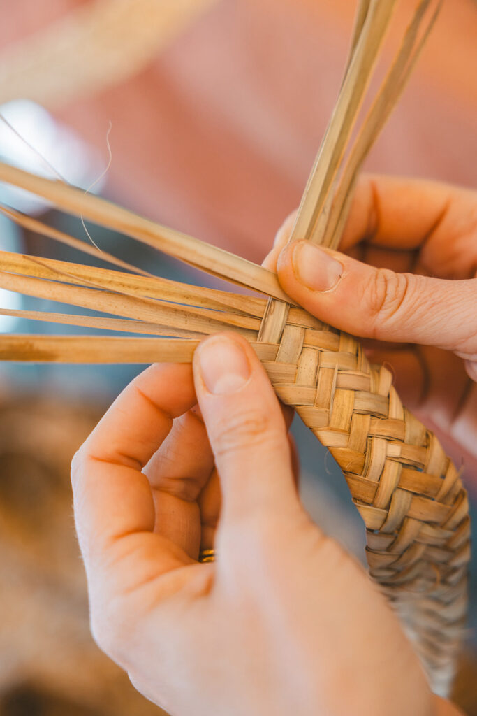 Mãos entrelaçando fibras naturais na técnica tradicional de empreita de palma durante um workshop de cestaria. A trama detalhada destaca a precisão artesanal e a herança do artesanato algarvio.