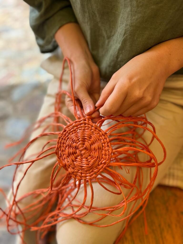Mãos tecendo uma peça em técnica de twining com fibras vermelhas durante um workshop de cestaria. A estrutura circular começa a ganhar forma, destacando o trabalho artesanal e a tradição da cestaria.